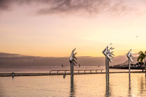 Cairns Esplanade Pool At Sunrise. Silver Fish Monuments Stand On Pillars Coming Out Of The Pool.